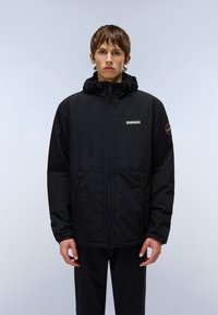 Ung man står framåt med en svart jacka med huva, dragkedja framtill och logotyp på vänster bröst och övre ärm.