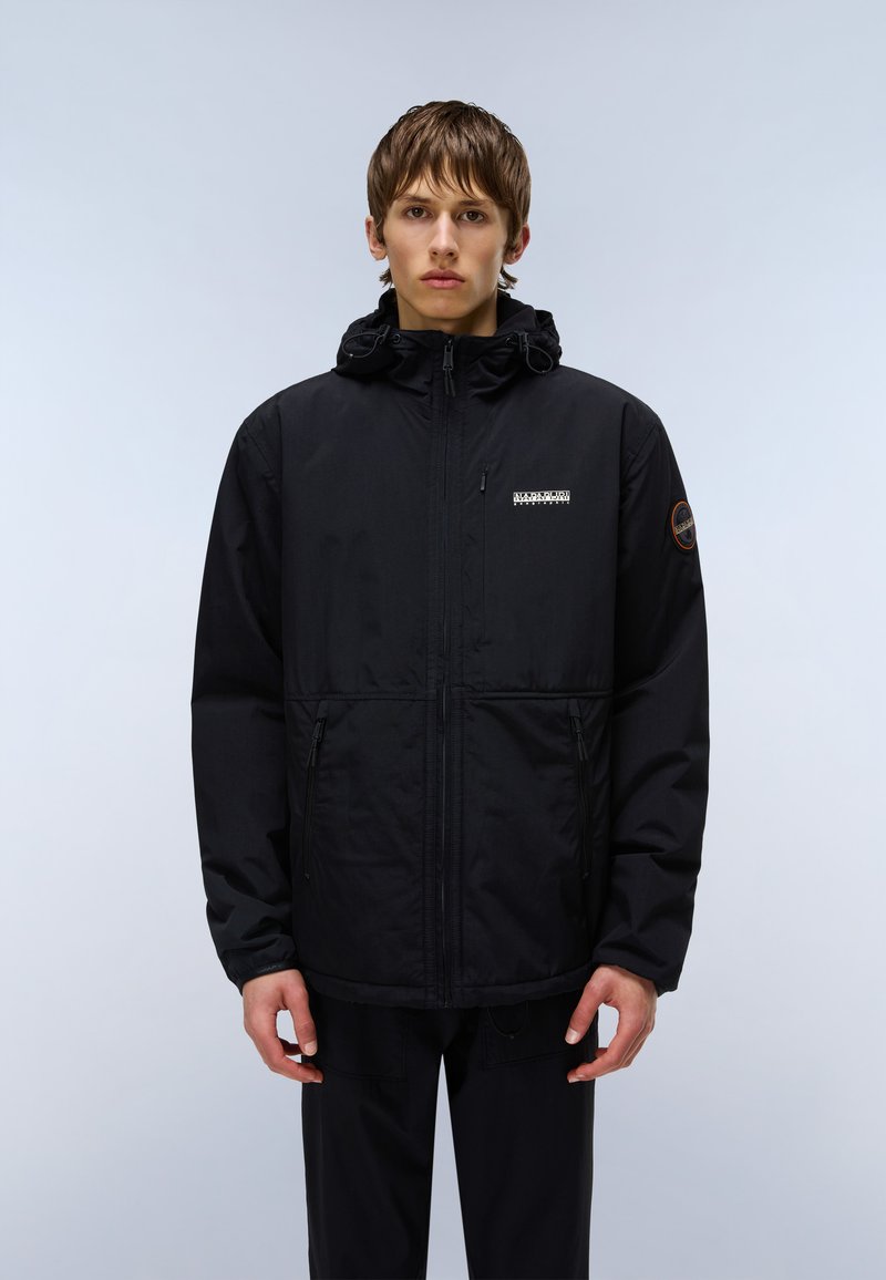 Ung man står framåt med en svart jacka med huva, dragkedja framtill och logotyp på vänster bröst och övre ärm.