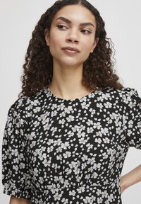 Robe noire avec un motif floral comprenant des fleurs blanches, col rond et manches bouffantes. Le tissu semble léger avec une texture douce.