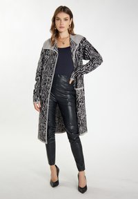 Langer grauer und schwarzer gemusterter Cardigan mit Kragen, offenem Front und Taschen, kombiniert mit schwarzen Kunstlederhosen und schwarzen High Heels.