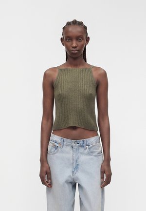 Donna con capelli intrecciati che indossa un top corto senza maniche a coste verde oliva e jeans a vita alta azzurri, in piedi davanti a uno sfondo bianco.