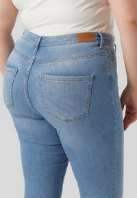 Lyseblå jeans laget av denim, med fem lommer, en brun lærpatch bak, og en glatt, noe elastisk tekstur.
