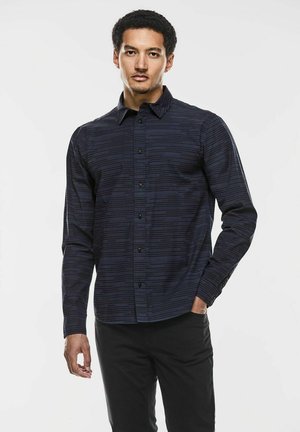 Marineblå langærmet skjorte med knaplukning, der har et horisontalt stribemønster, spidse kraver og sorte knapper, stylet med sorte bukser.