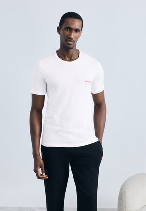 Weißes Baumwoll-T-Shirt mit rundem Halsausschnitt und kurzen Ärmeln, mit kleinem rotem "HUGO"-Logo auf der Brust; kombiniert mit schwarzen, strukturierten Hosen.