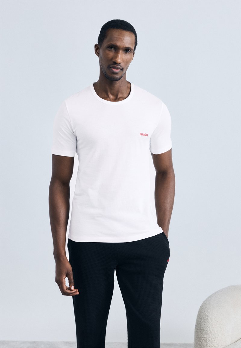 Baltas medvilninis marškinėlis su apvalia iškirpte ir trumpomis rankovėmis, ant krūtinės mažas raudonas „HUGO“ logotipas; dera su juodomis tekstūruotomis kelnėmis.