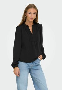 Svart blus med lång ärm, V-ringning och rynkig krage. Strukturerad tyg, dekorativ sömnad fram och samlade manschetter.