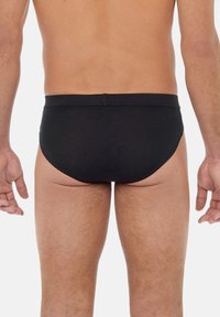 Schwarze Baumwollslips mit glatter Oberfläche, die ein einfarbiges Design und einen elastischen Bund aufweisen, von hinten auf einem neutralen Hintergrund betrachtet.