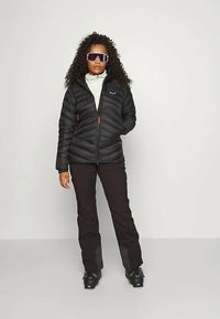 Γυναίκα με σγουρά μαλλιά, ντυμένη με μαύρο μπουφάν, λευκό ζιβάγκο, μαύρα παντελόνια, μπότες σκι και μεγάλα σκουρόχρωμα γυαλιά σκι, που στέκεται μπροστά σε απλή λευκή φόντο.