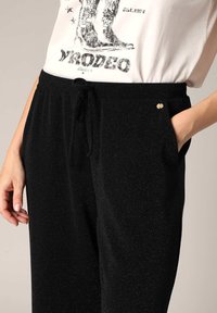 Zwarte joggingbroek gemaakt van zachte, gestructureerde stof met een subtiele glans. Voorzien van een koord in de taille en zijzakken. Kleine gouden accentdetails.