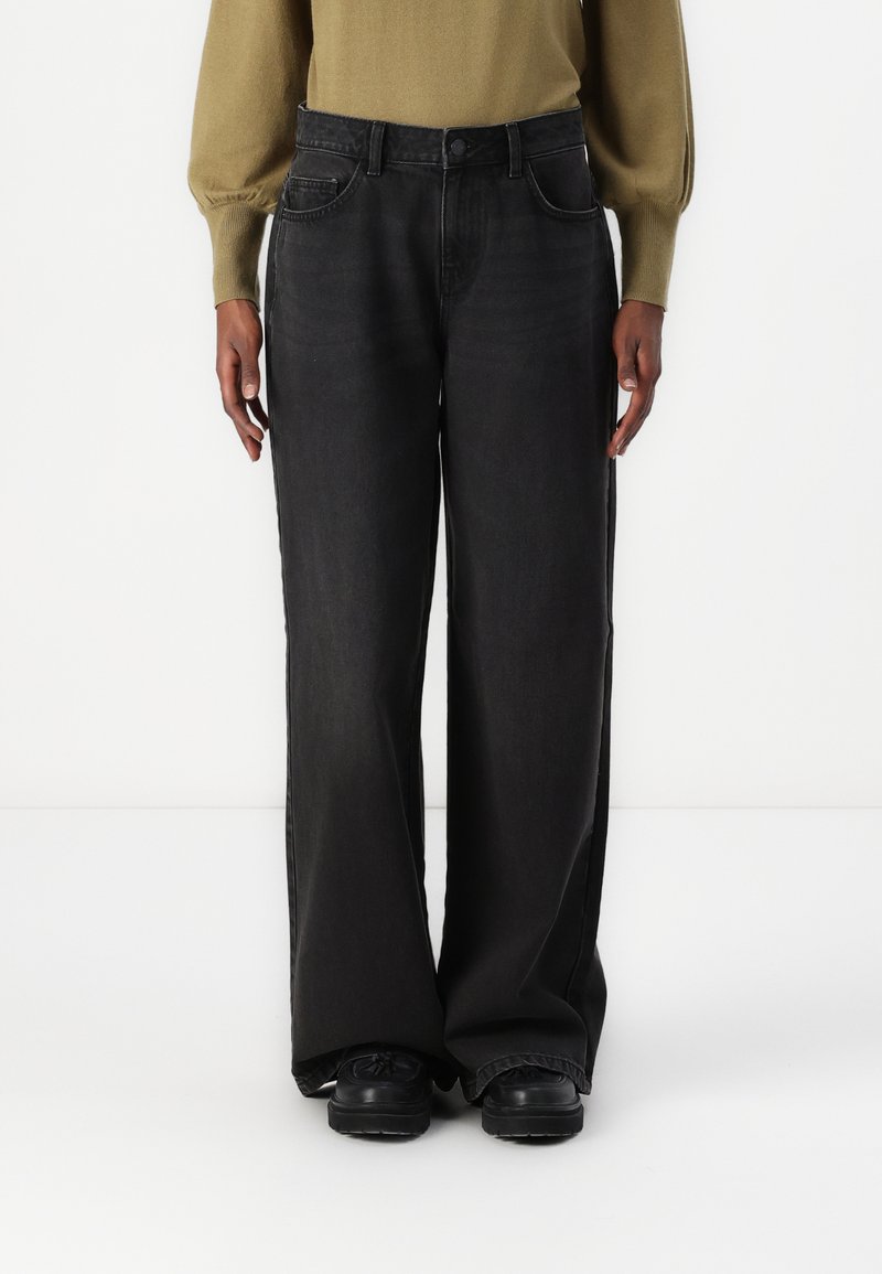 Jean en denim noir à jambes larges et taille haute, avec un design à cinq poches et un délavage subtil. Associé à un pull ajusté vert olive.