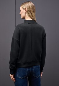 Sudadera negra de corte relajado, con puños acanalados y un cuello alto. Textura suave, con una costura vertical en la parte posterior. Llevada con jeans azules.