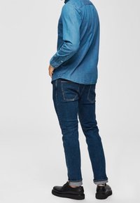 Muž oblečený v tmavě modrých džínech, odpovídající modré džínové košili a černých společenských botách, stojící se rukama blízko kapes.