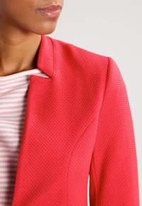 Rød struktureret blazer med en skarp krave. Under den, en lys stribet top. Fokus på stoffets mønster og blazerens strukturerede design.