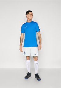 Modrá sportovní košile s krátkými rukávy a kulatým výstřihem; bílé kraťasy s logem; černé boty; bílé ponožky s černými detaily.