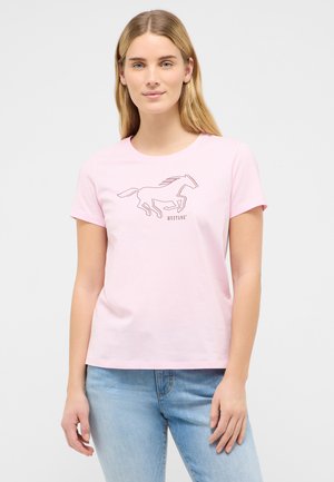 Junge Frau mit langen blonden Haaren, die ein hellrosa T-Shirt mit einer Kontur eines laufenden Pferdes und dem Schriftzug „MUSTANG“ trägt, kombiniert mit hellblauen Jeans.