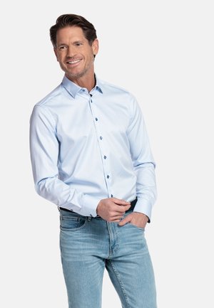 Lachende man die een lichtblauw overhemd met knopen en een spijkerbroek draagt, staand met één hand in zijn zak en de andere hand die zijn overhemdmanchet aanpast.