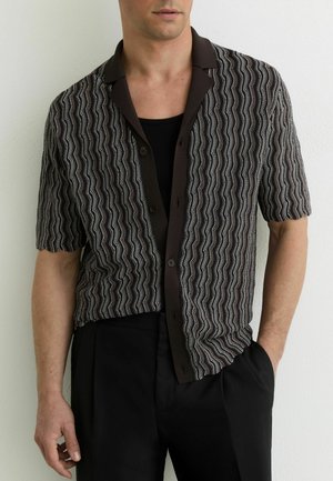 Mand iført en kortærmet skjorte med bølgete sorte og hvide striber over en sort tanktop, sorte bukser, hånd i lommen.