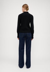 Maglione nero in maglia con colletto alto, abbinato a jeans in denim blu scuro a vestibilità ampia. Il tessuto è liscio con dettagli minimi sulle tasche.