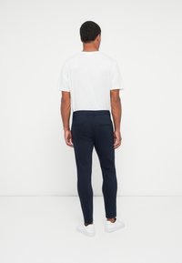 Pantaloni slim-fit blu navy con una texture liscia, due tasche posteriori e passanti per la cintura, abbinati a una t-shirt bianca a maniche corte e sneakers bianche.