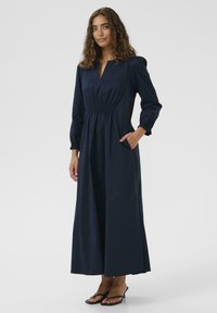 Marineblaues langes Kleid mit V-Ausschnitt, gerafftem Taillengummi und langen Ärmeln. Hergestellt aus leichtem Stoff und mit seitlichen Taschen.