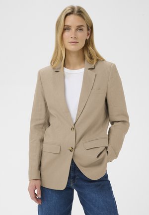Woman with blonde hair wearing a beige blazer over a white shirt and blue jeans, standing against a plain light background.