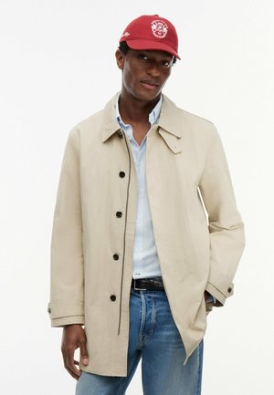 Mann trägt einen beigen, geknöpften Mantel, hellblau gestreiftes Hemd, blaue Jeans, schwarzen Gürtel und eine rote Kappe mit weißem Emblem, steht mit einer Hand in der Tasche.