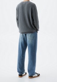 Grauer, gestrickter Pullover mit lockerer Passform, kombiniert mit hellblauen, weit geschnittenen Jeans. Das Schuhwerk hat schwarze und weiße Akzente.