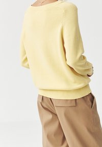Żółty sweter dzianinowy z długimi rękawami i szerokim dekoltem. Tekstura jest gładka, z prążkowanymi mankietami i dołem. Noszony z beżowymi spodniami.