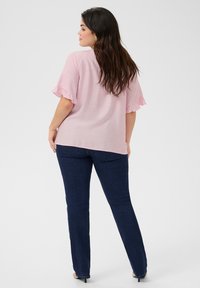 Blusa rosa con maniche corte arricciate, abbinata a jeans dritti blu scuro. Il tessuto sembra leggero e strutturato.