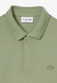 Polo shirt verde chiaro con colletto e un piccolo logo di coccodrillo ricamato sul lato sinistro del petto, etichettato "Lacoste Regular Fit."