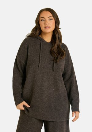 Femme portant un hoodie tricoté marron foncé oversize et un pantalon assorti, debout devant un fond clair uni.