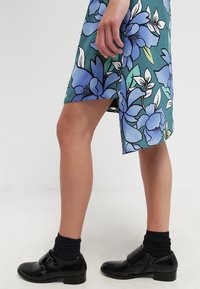 Jambes portant une robe bleue fleurie avec une fente latérale, des chaussettes noires et des chaussures noires brillantes à boucle. Une main repose sur le côté.