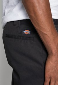 Detailní záběr na osobu v černých kalhotách Dickies s logem na kapsičce nad zadní kapsou a bílou košilí zastrčenou uvnitř.