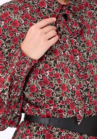 Blouse florale en tissu noir avec un motif de fleurs rouges et vertes, manches smockées et un détail à nouer au cou. Fini avec une ceinture noire.