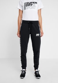 Svarta sweatpants med dragsko i midjan, sidotryck med logotyp och avslutade anklar, kombinerade med en vit grafisk t-shirt. Bärs med svarta sneakers.