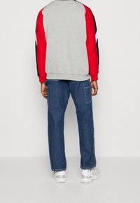 Felpa grigia con maniche rosse e blu navy, abbinata a jeans di denim blu ampi. Presenta una vestibilità classica e polsini e orlo a costine. Sneakers bianche.