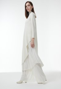 Lang witte jurk met een hoge hals, vloeiende gelaagde ontwerp en gestructureerde stof, gecombineerd met witte enkellaarzen.