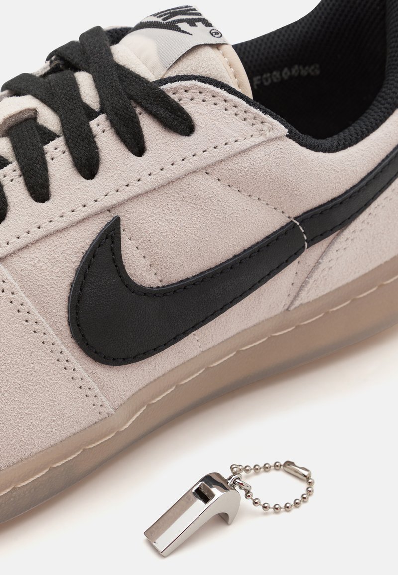 Sneaker in camoscio grigio chiaro con accenti neri, lacci neri e suola in gomma traslucida; presenta un piccolo fischietto argentato attaccato.