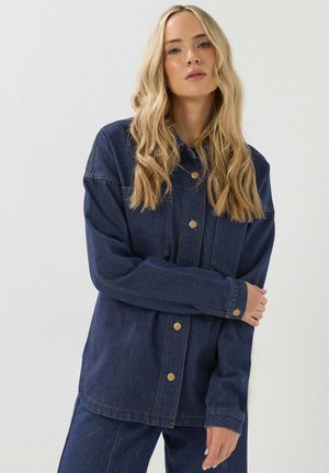 Vrouw met lang blond haar die een donkerblauwe spijkerjas en bijpassende broek draagt, staand met één arm over haar borst gekruist.