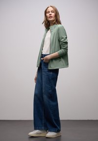 Giacca verde chiaro con zip sopra una maglietta bianca, abbinata a jeans a gamba larga blu e sneakers bianche; tessuto liscio, vestibilità rilassata e stile casual.