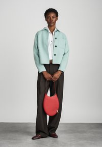 Giacca di lana verde menta, camicia bianca, pantaloni marroni, borsa curva rossa e scarpe scure. Concentrati sul contrasto dei colori e sui dettagli dei materiali.