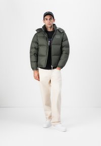 Giacca verde oliva imbottita con chiusura a zip e bottoni a pressione, caratterizzata da un collo alto e due tasche laterali. Il modello indossa pantaloni beige chiaro.