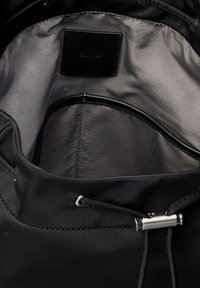 Interno di una borsa in nylon nera con fodera grigia liscia, un piccolo etichetta in pelle nera e un cordoncino nero con un fermaglio cilindrico in metallo argentato.