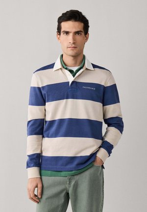 Joven con una camiseta de rugby de manga larga a rayas beige y azul con cuello blanco, camiseta interior verde y pantalones verdes, de pie en posición neutral.