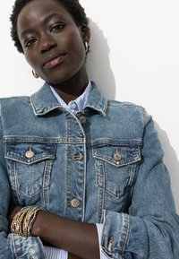 Veste en denim avec deux poches poitrine, fermeture à boutons et couleur bleu délavé. Superposée sur une chemise à rayures bleu clair et blanche.