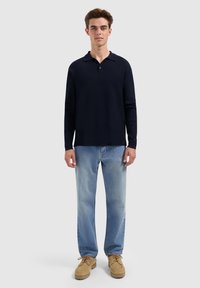 Marineblauwe polo met lange mouwen, een kraag en knoopdetails, gecombineerd met lichtblauwe denim jeans en beige moccasins.