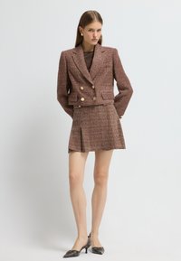 Blazer de tweed marrón con botones dorados dobles, combinado con una mini falda plisada a juego. Modelo con tacones negros de punta afilada.