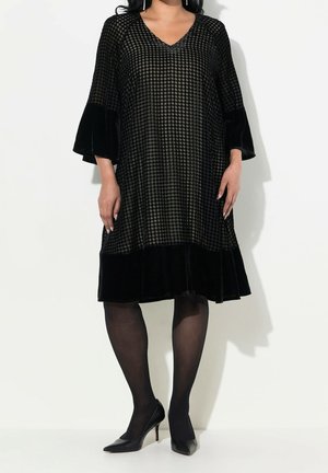 Frau trägt ein knielanges schwarzes Samtkleid mit kariertem Muster, Glockenärmeln, schwarzen Strumpfhosen und schwarzen Schuhen mit Absatz, steht vor weißem Hintergrund.