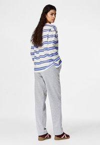 Woman with long hair wearing a white, blue, and black striped long-sleeve shirt, gray pants, and burgundy sneakers, standing with hands in pockets.