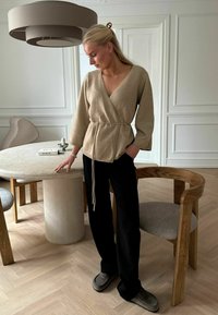 Beige Wickeloberteil mit langen Ärmeln und einem Knoten in der Taille, kombiniert mit schwarzen Hosen und grauen Slippern, in einer modernen Inneneinrichtung.